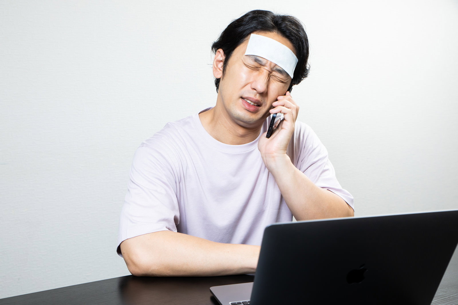 冷却シートを額に貼りスマートフォンで通話をする白いシャツの男性［モデル：大川竜弥］