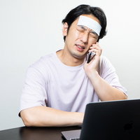 冷えピタを額に貼って通話する発熱中の男性の写真