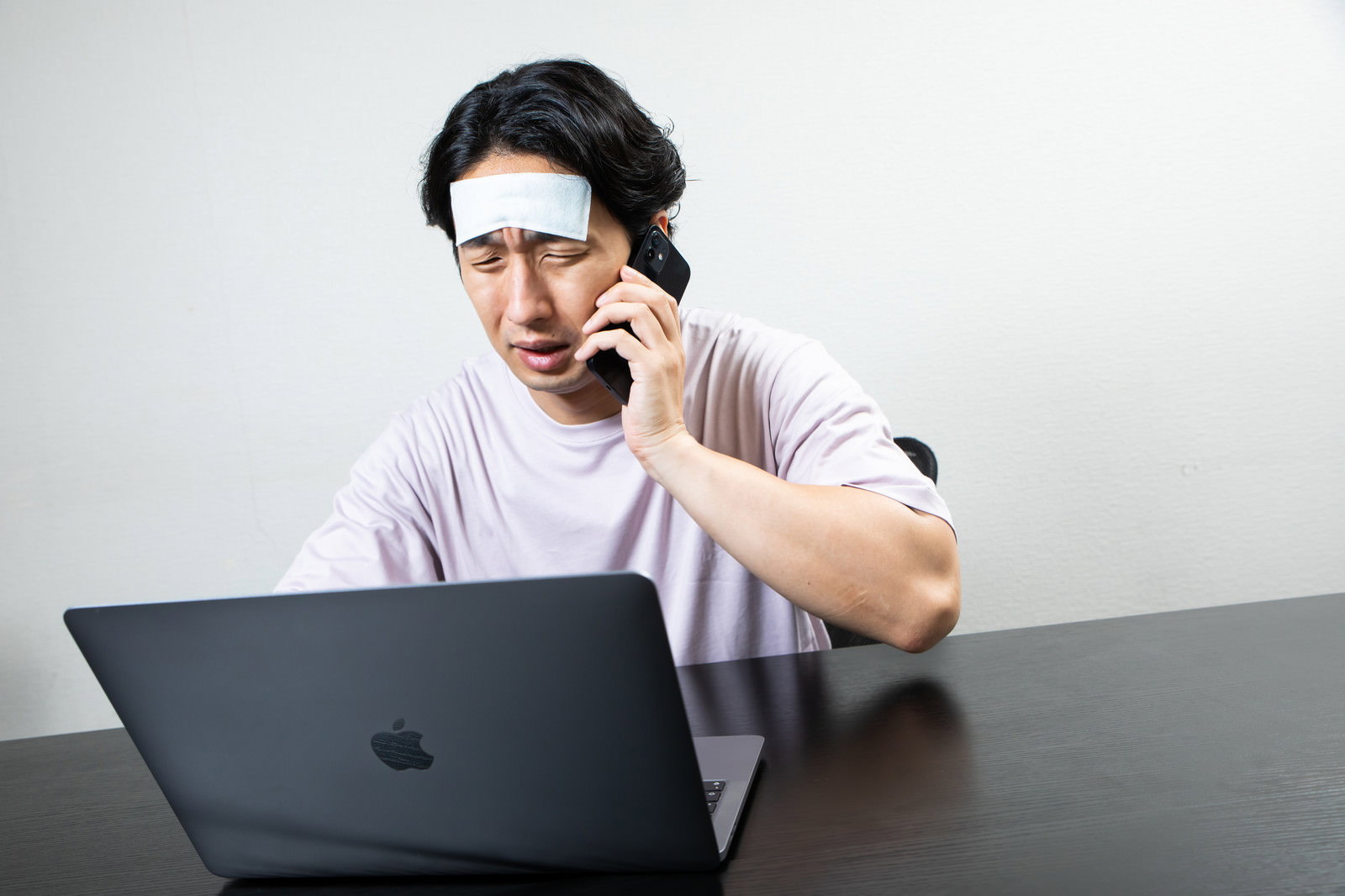 冷えピタを額に貼りながらノートパソコンで作業し電話をする男性［モデル：大川竜弥］