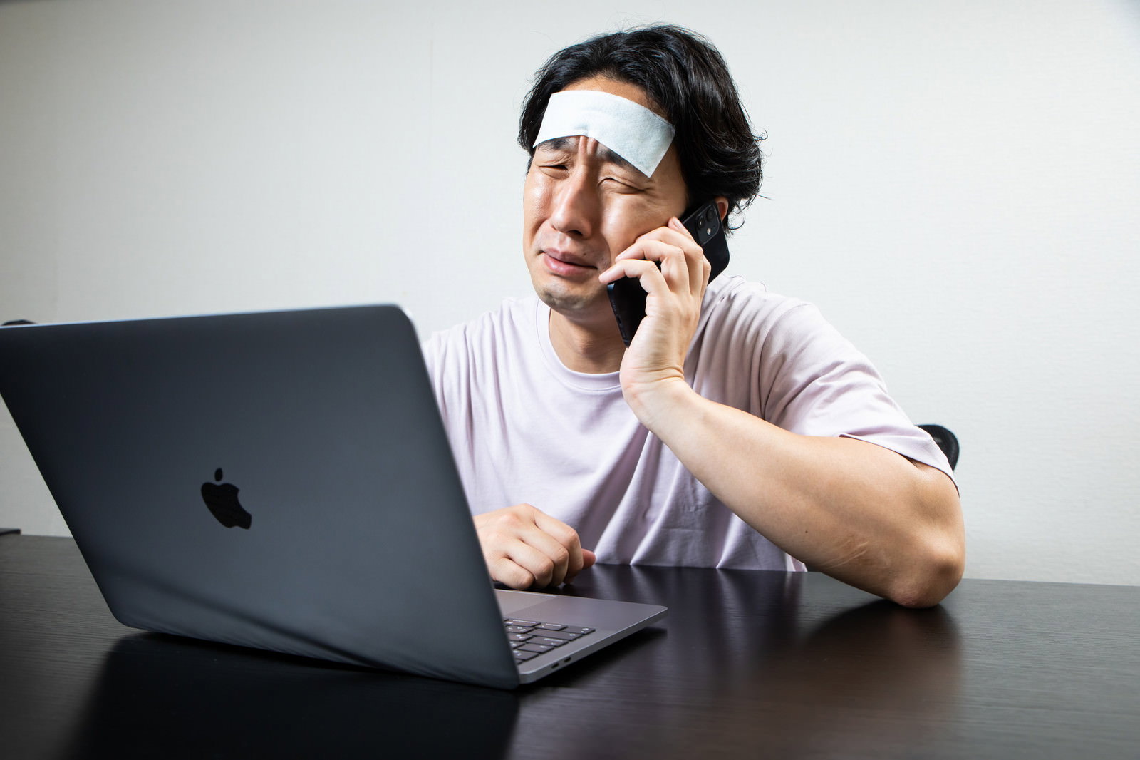 額に冷えピタを貼った男性が在宅でノートパソコンを使いながら電話をしている［モデル：大川竜弥］