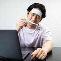 額に冷えピタを貼った体温計を見つめて仕事が手につかない男性の写真