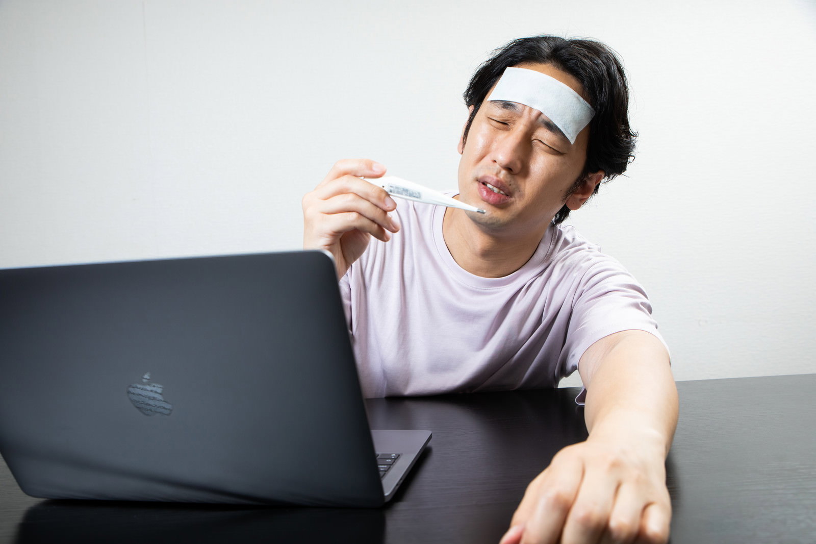 体温計を手に持ち冷却シートを額に貼ってうなだれる在宅勤務中の男性［モデル：大川竜弥］