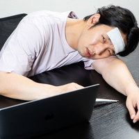 仕事中に机でぐったりして体温計を持つ高熱男子の写真