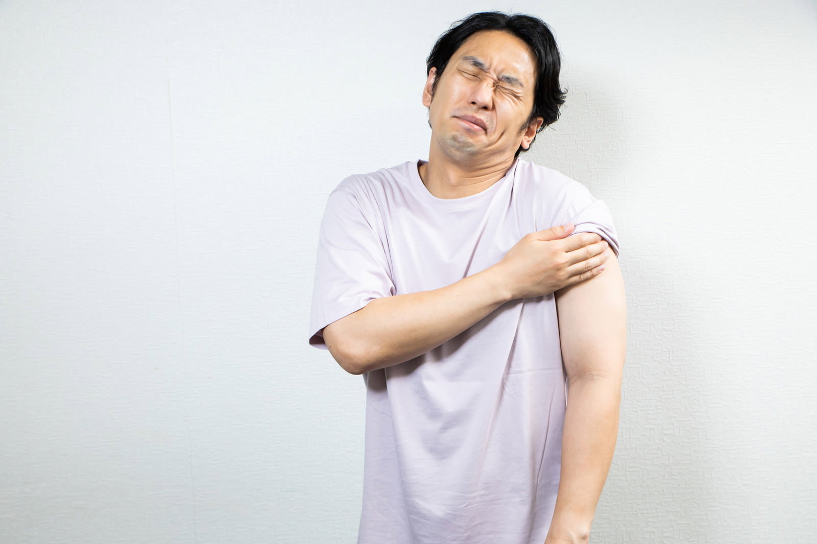 ワクチン接種後に左腕を抑えて困った表情を見せる白いTシャツの男性［モデル：大川竜弥］