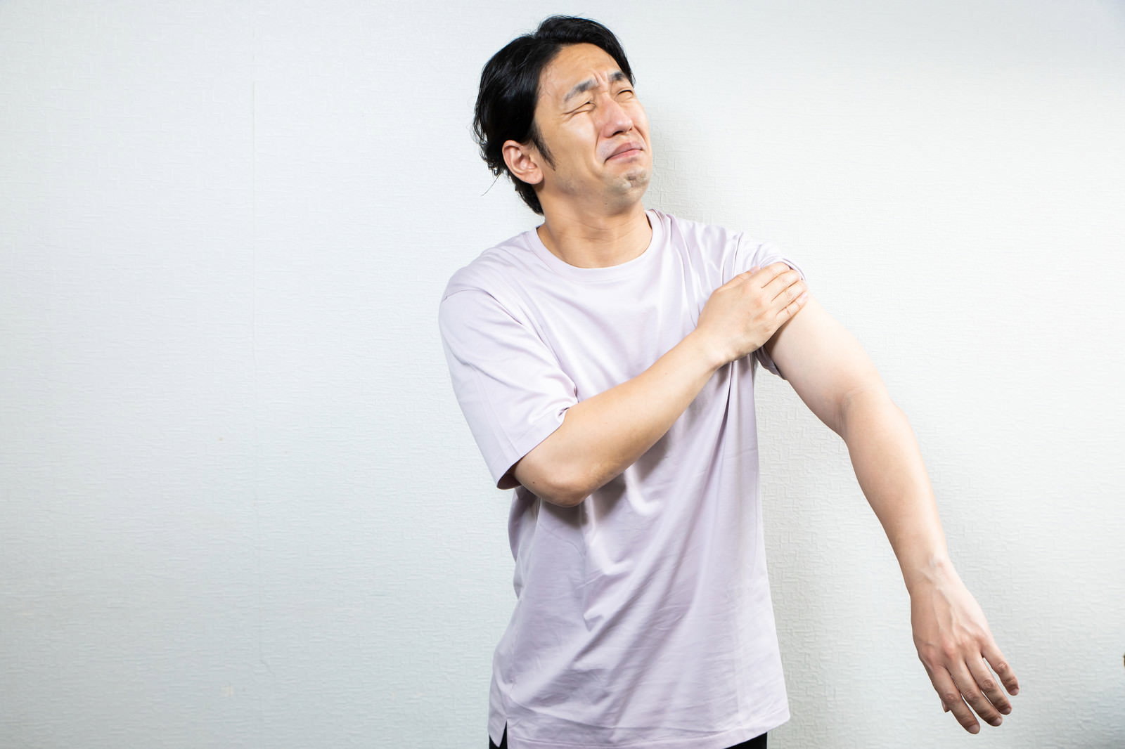 左腕を抑えて痛そうな表情でしかめっ面をする白いTシャツの男性［モデル：大川竜弥］