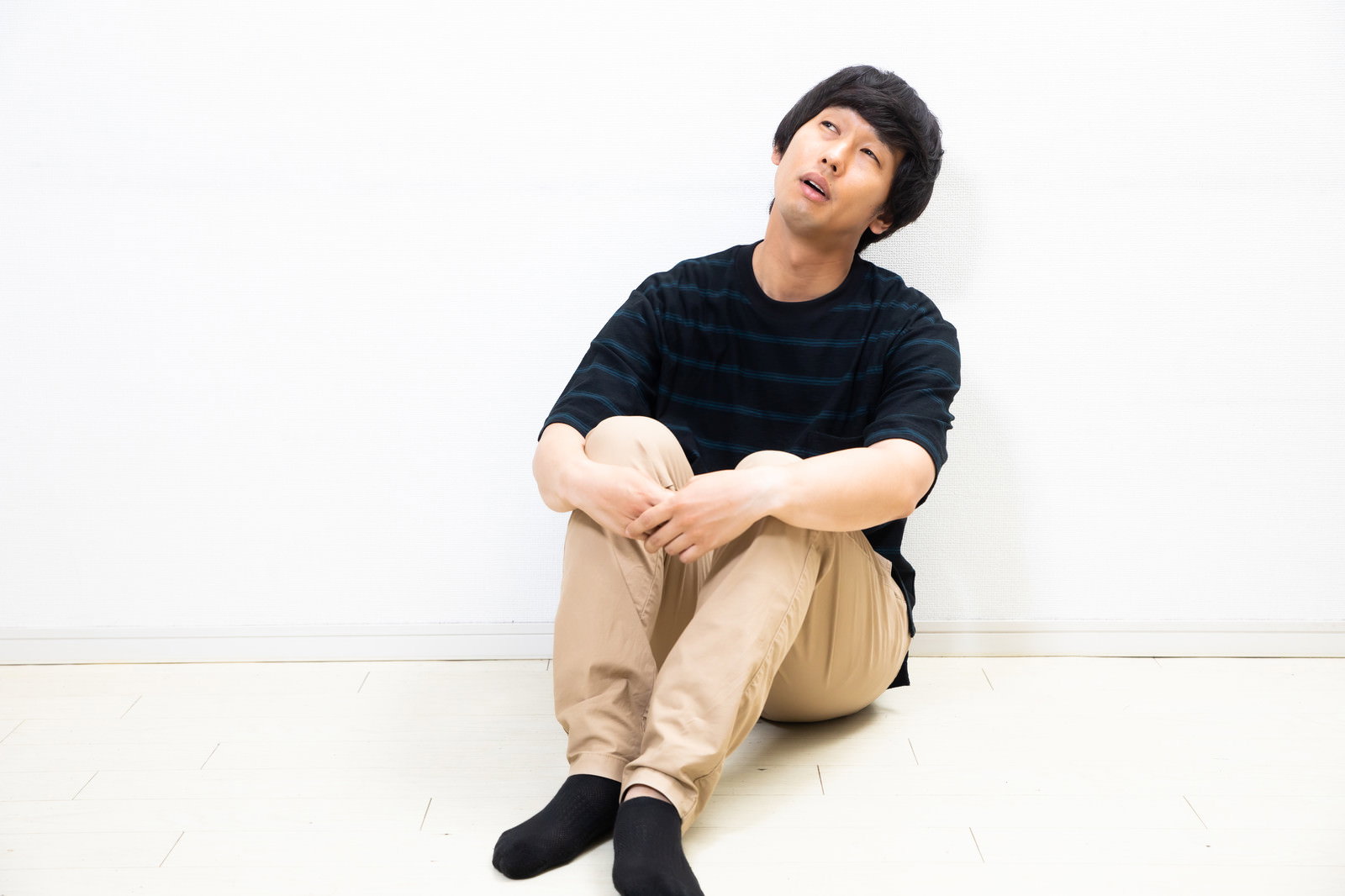 黒い服を着た男性が床に体育座りで座り、膝を抱えながらボーっとしている［モデル：大川竜弥］