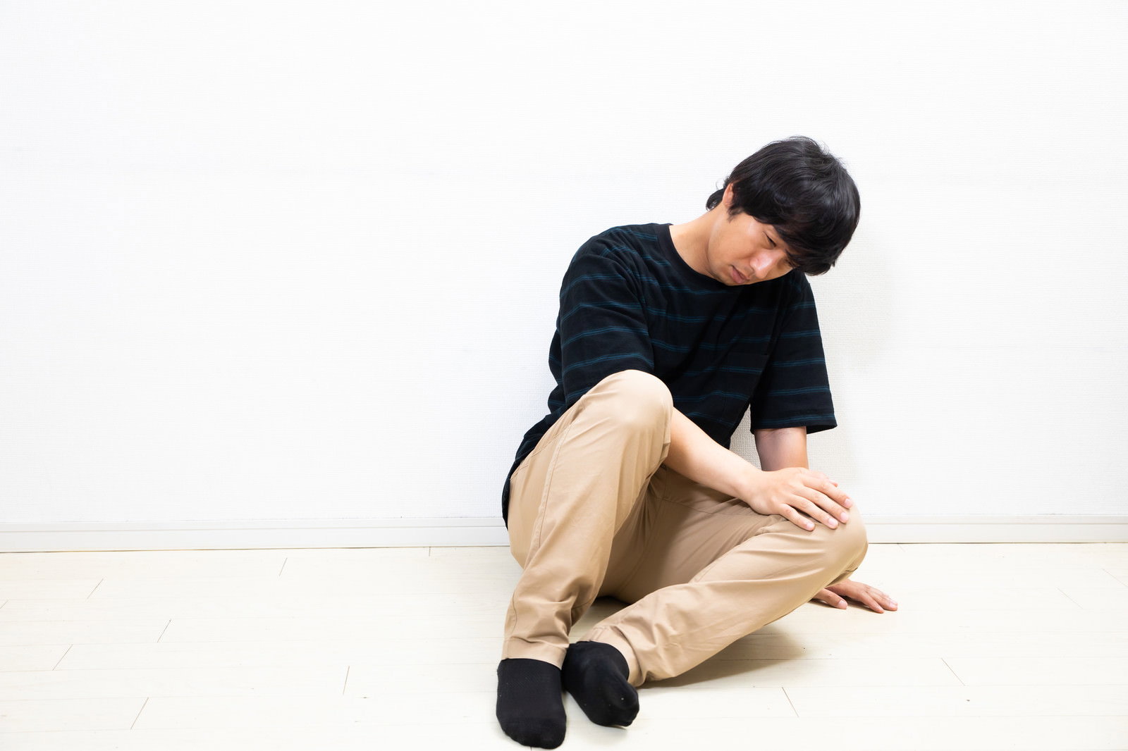 黒いトップスとベージュのパンツを着た男性が座り込んでうずくまっている様子［モデル：大川竜弥］