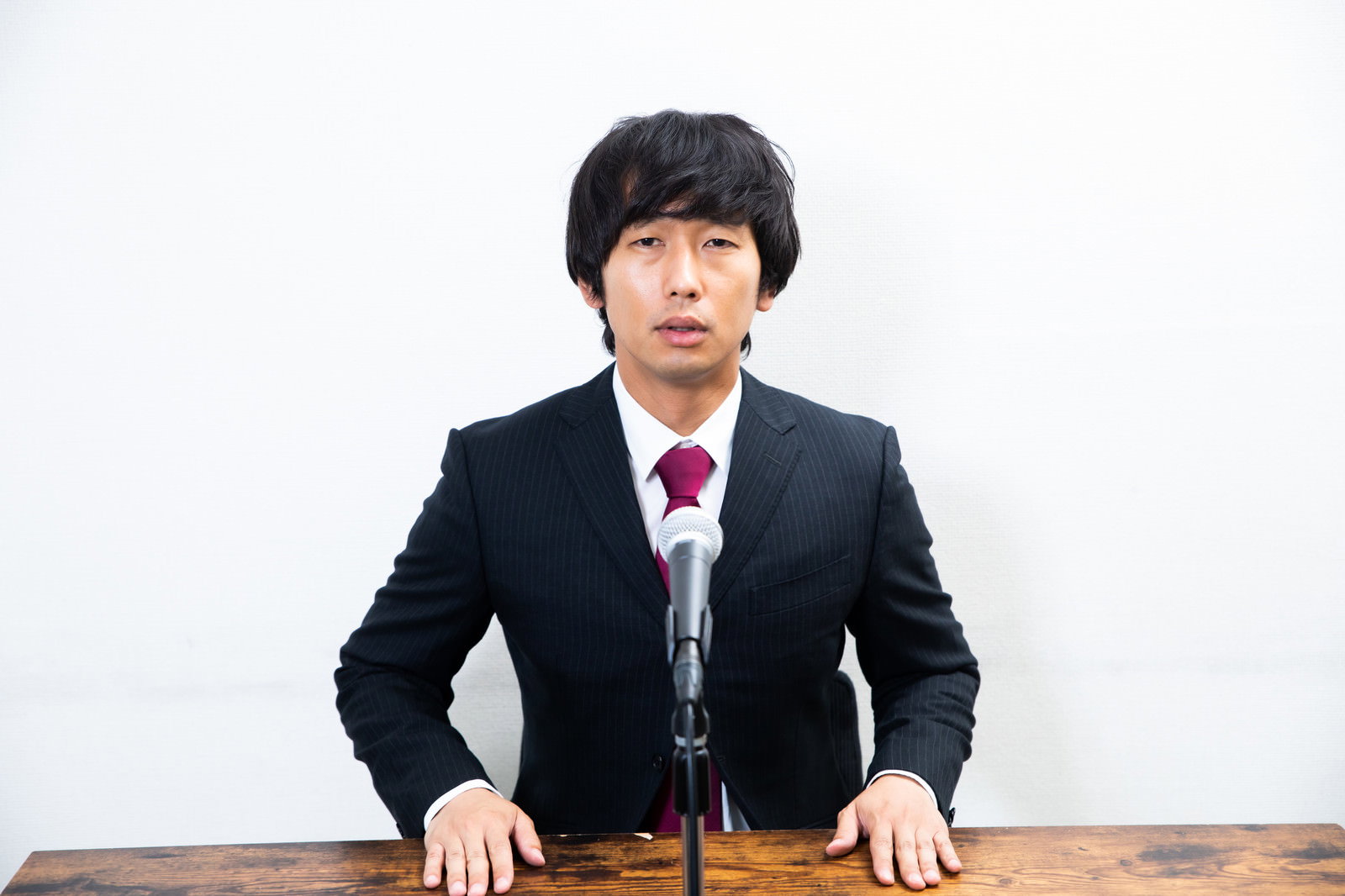 黒いスーツを着た男性が演壇のマイクの前で謝罪している［モデル：大川竜弥］