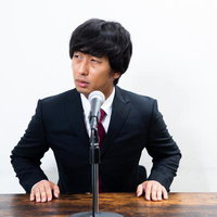 マイクの前で横を向く記者会見中の男性の写真