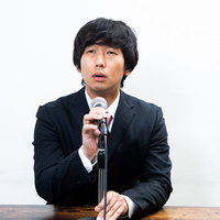 記者会見中にマイク位置を調整する黒いスーツの男性の写真