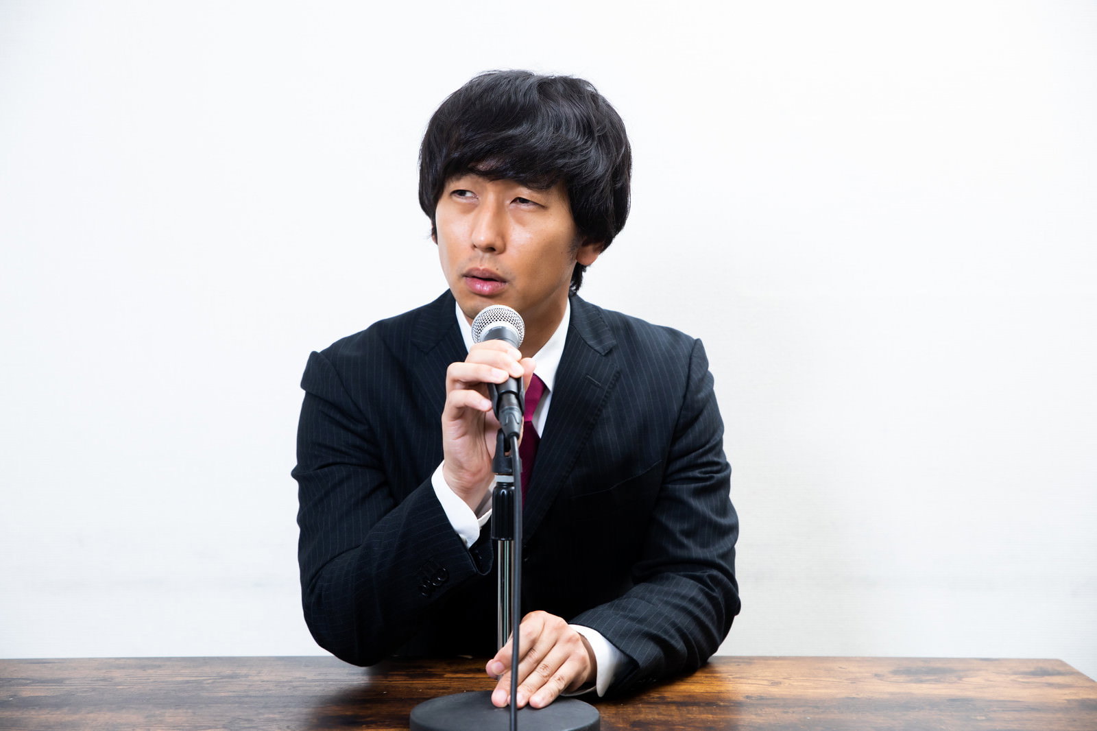 黒いスーツを着た男性がテーブルに座りマイクを持って会見している［モデル：大川竜弥］