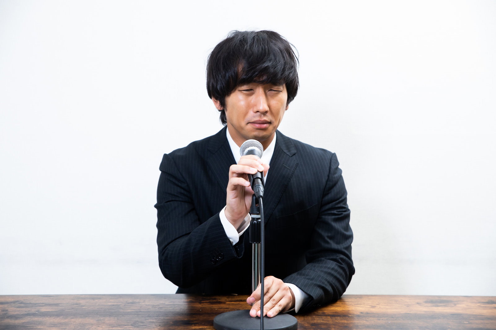 謝罪会見で困った表情を浮かべマイクの前に座る黒いスーツの男性［モデル：大川竜弥］