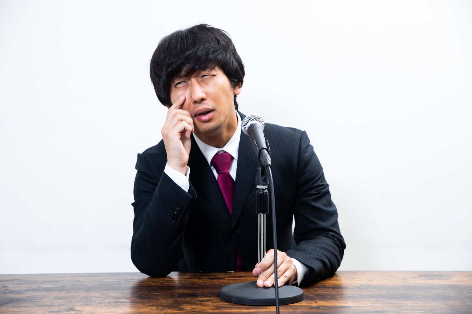 手を額に当てて困った表情でマイクの前に座る男性［モデル：大川竜弥］