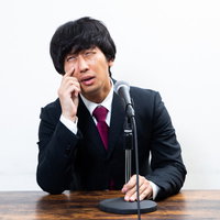 スーツ姿で謝罪する男性の記者会見、手を額に当てて困惑する表情の写真