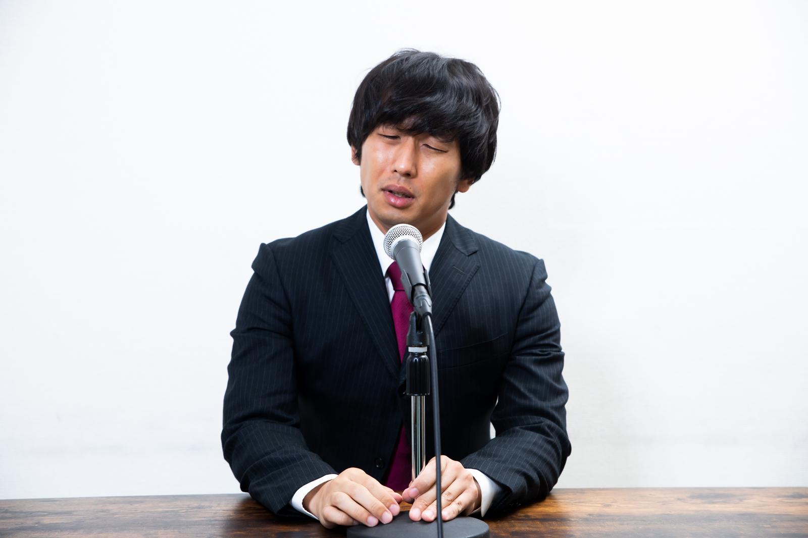 マイクの前で話す黒いスーツ姿の男性［モデル：大川竜弥］