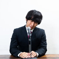 謝罪会見で涙を流し頭を下げる黒いスーツ姿の男性の写真