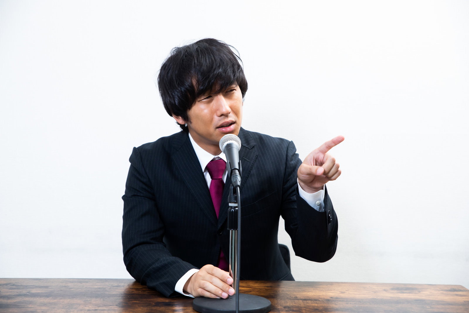 記者会見で右手の指を向けながら発言するスーツを着た男性［モデル：大川竜弥］