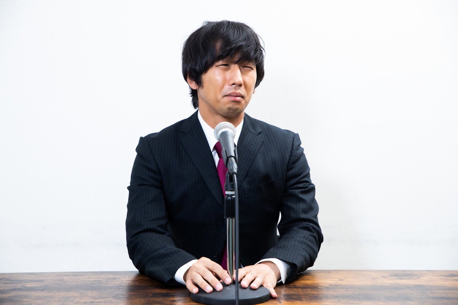 黒いスーツを着て苦渋の表情でマイクの前に座る男性