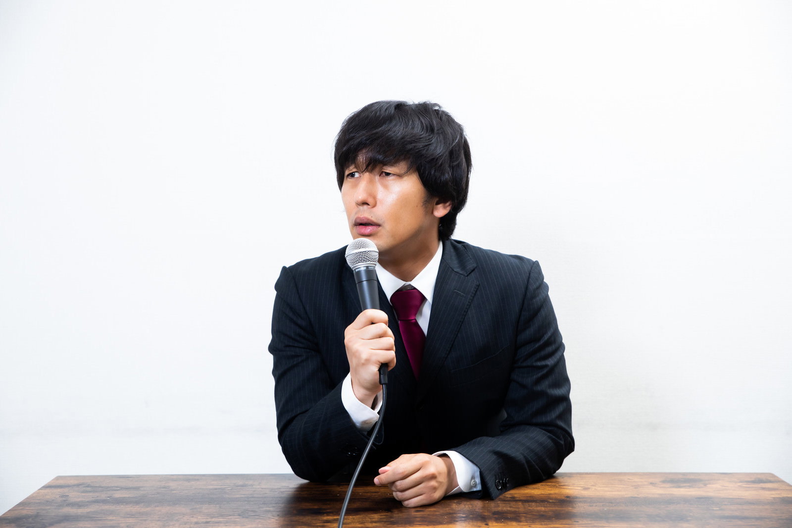 木製テーブルの前でマイクに向かって真剣な表情を見せる黒いスーツの男性［モデル：大川竜弥］