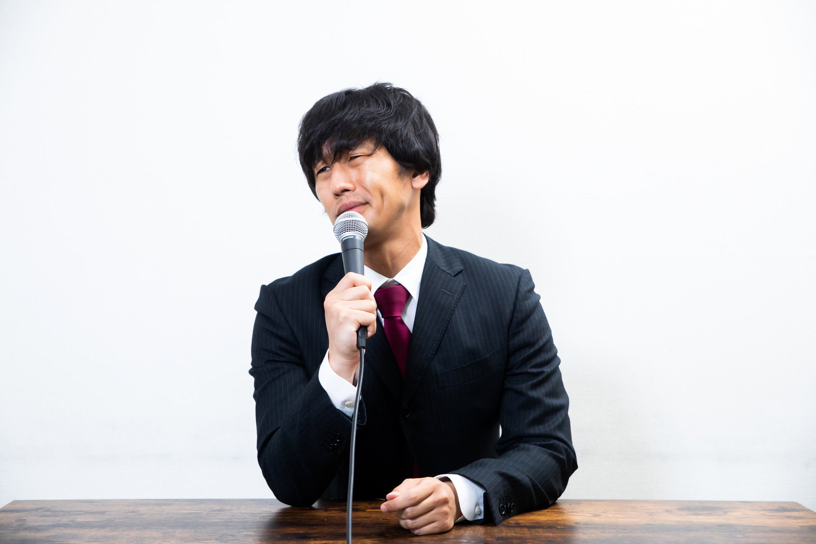 黒いスーツを着た男性が机に向かってしかめっ面で会見している様子［モデル：大川竜弥］