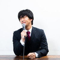 黒いスーツを着てしかめっ面で会見する男性の写真