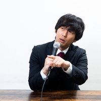 会見中に質問した記者を煽る黒いスーツ姿の男性の写真