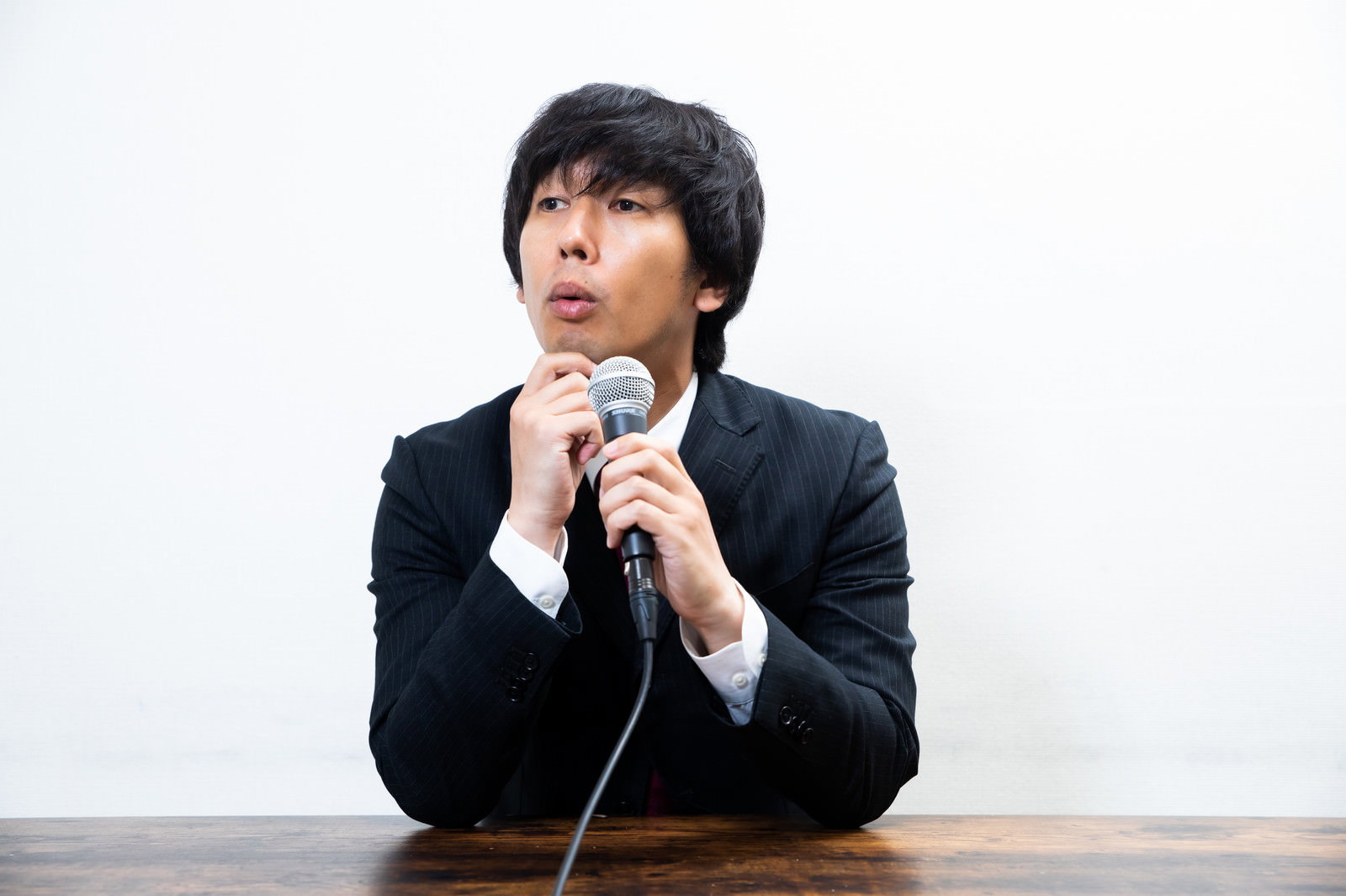 記者会見でマイクを前に手をあごに当てて考える表情のスーツ姿の男性［モデル：大川竜弥］