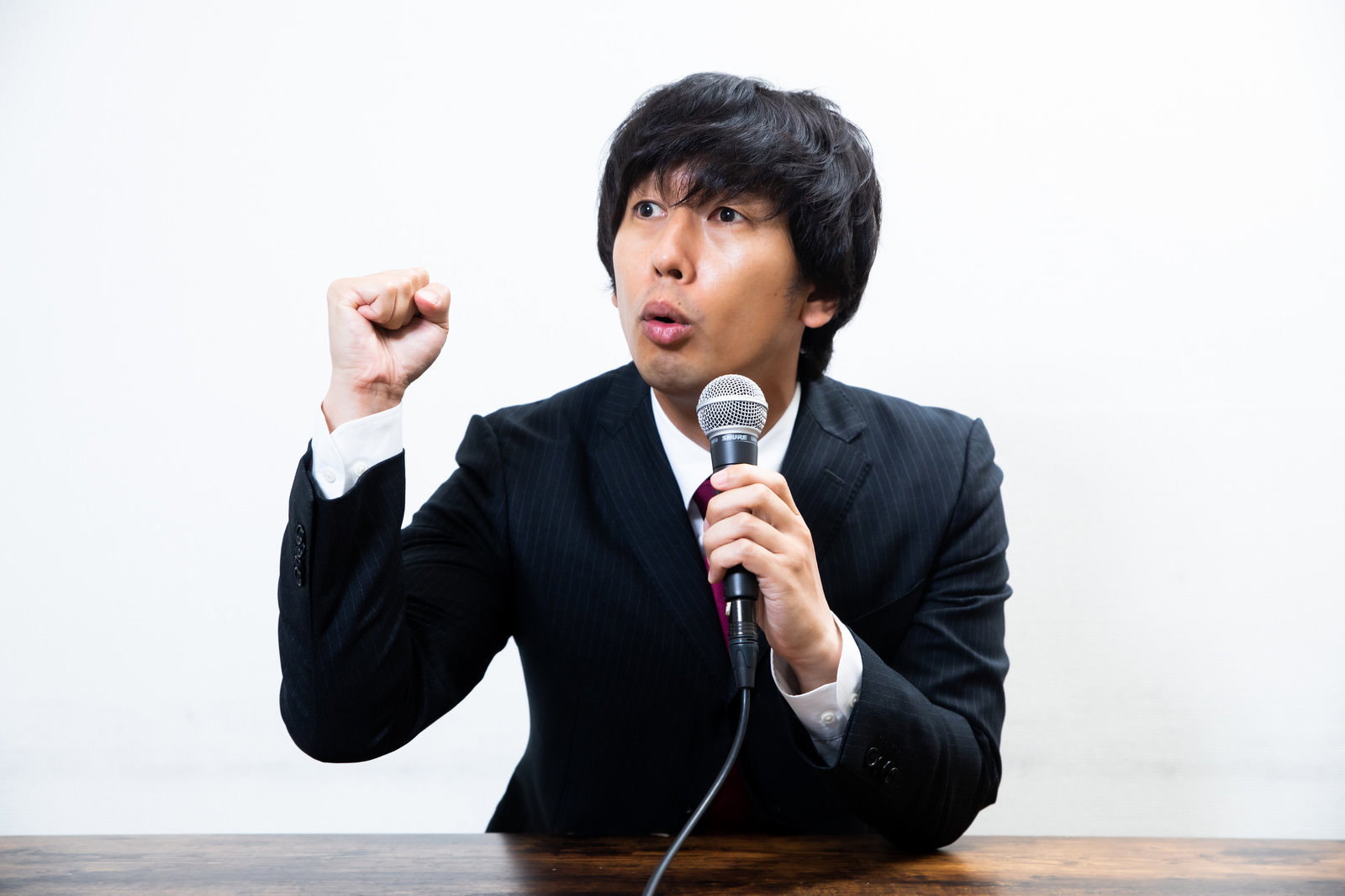 黒いスーツを着た男性がマイクを持ち拳を握りしめながら会見している［モデル：大川竜弥］