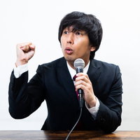 黒いスーツで拳を握りしめながら会見する男性の写真