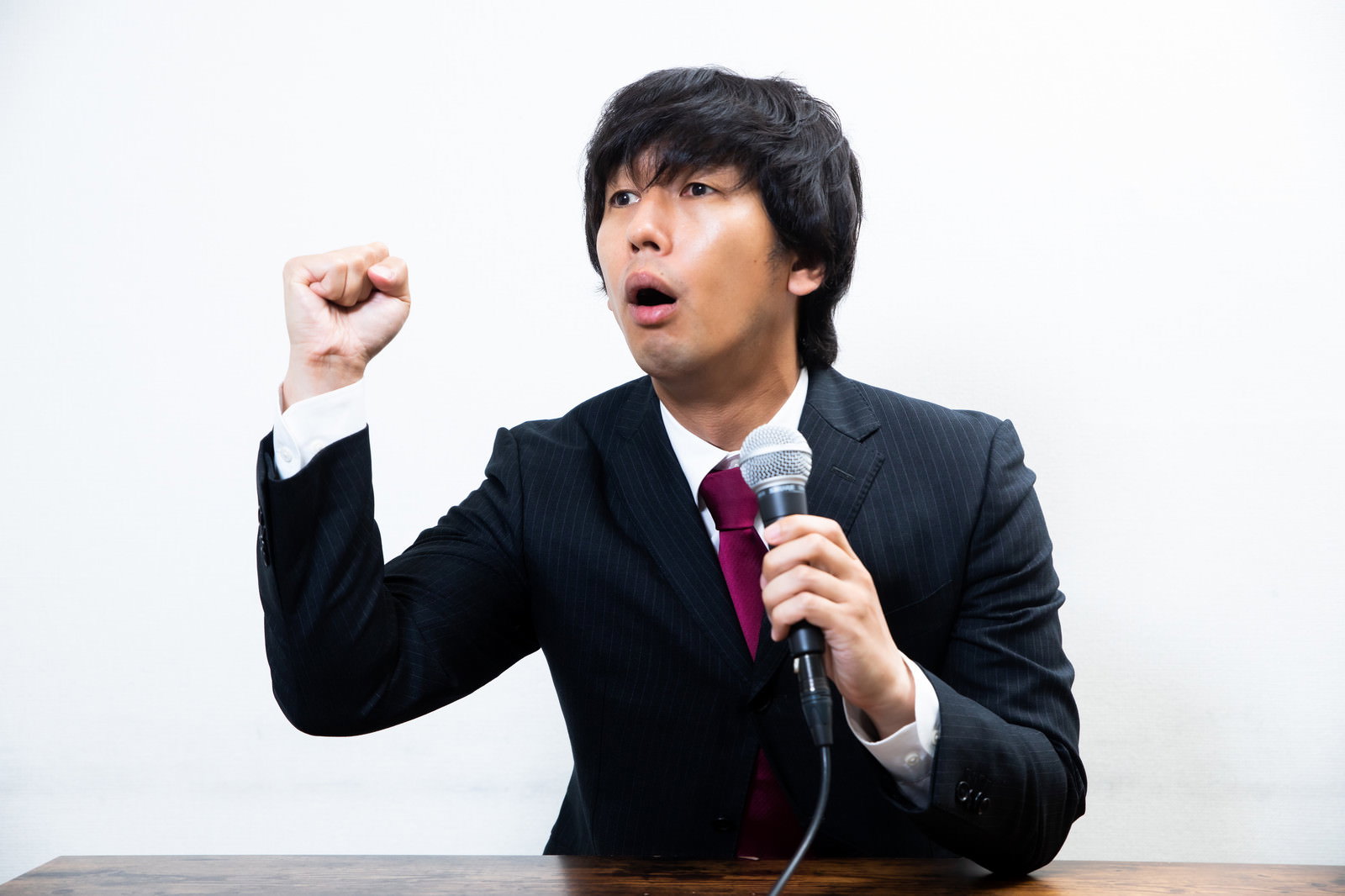マイクを持ち右手を上げて力説するスーツ姿の男性［モデル：大川竜弥］