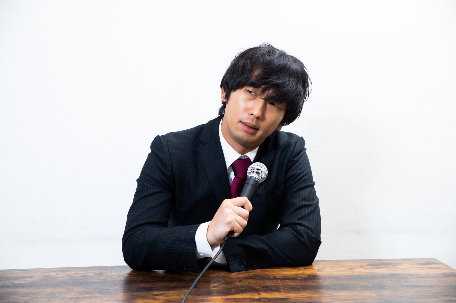 記者会見でマイクを持ち無愛想な表情で話すスーツ姿の男性［モデル：大川竜弥］