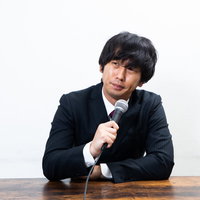 記者会見で無愛想にマイクを持つ男性の写真