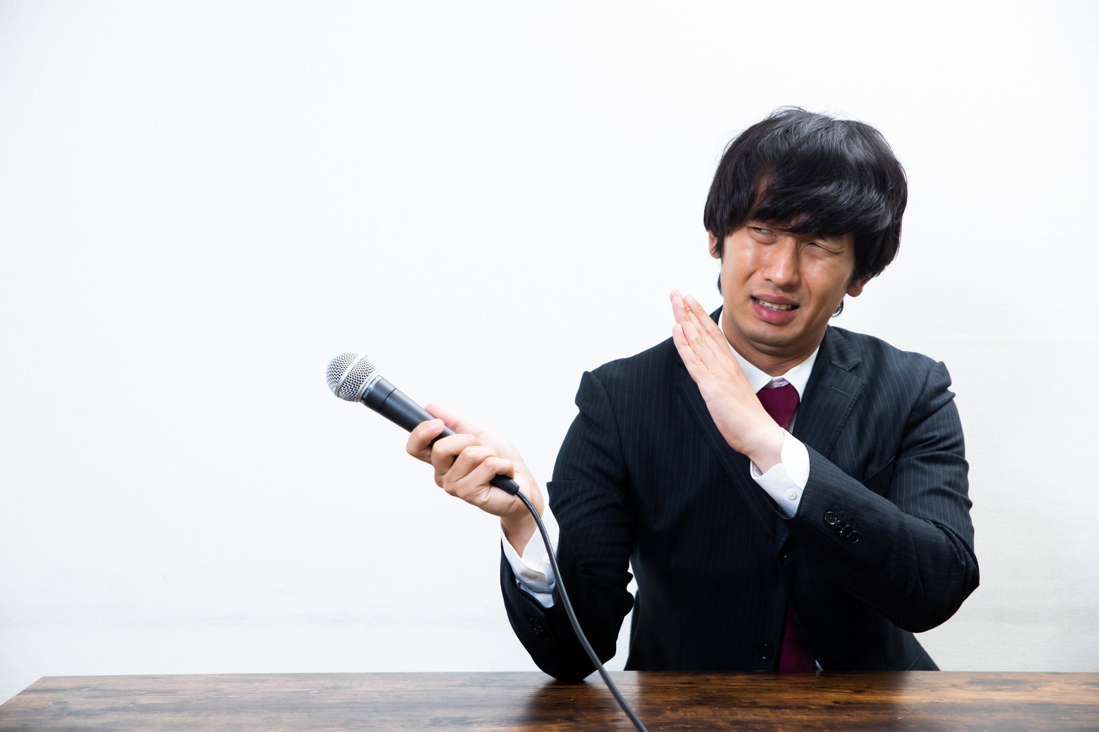 黒いスーツを着た男性が机に座り困った表情でマイクを持っている［モデル：大川竜弥］