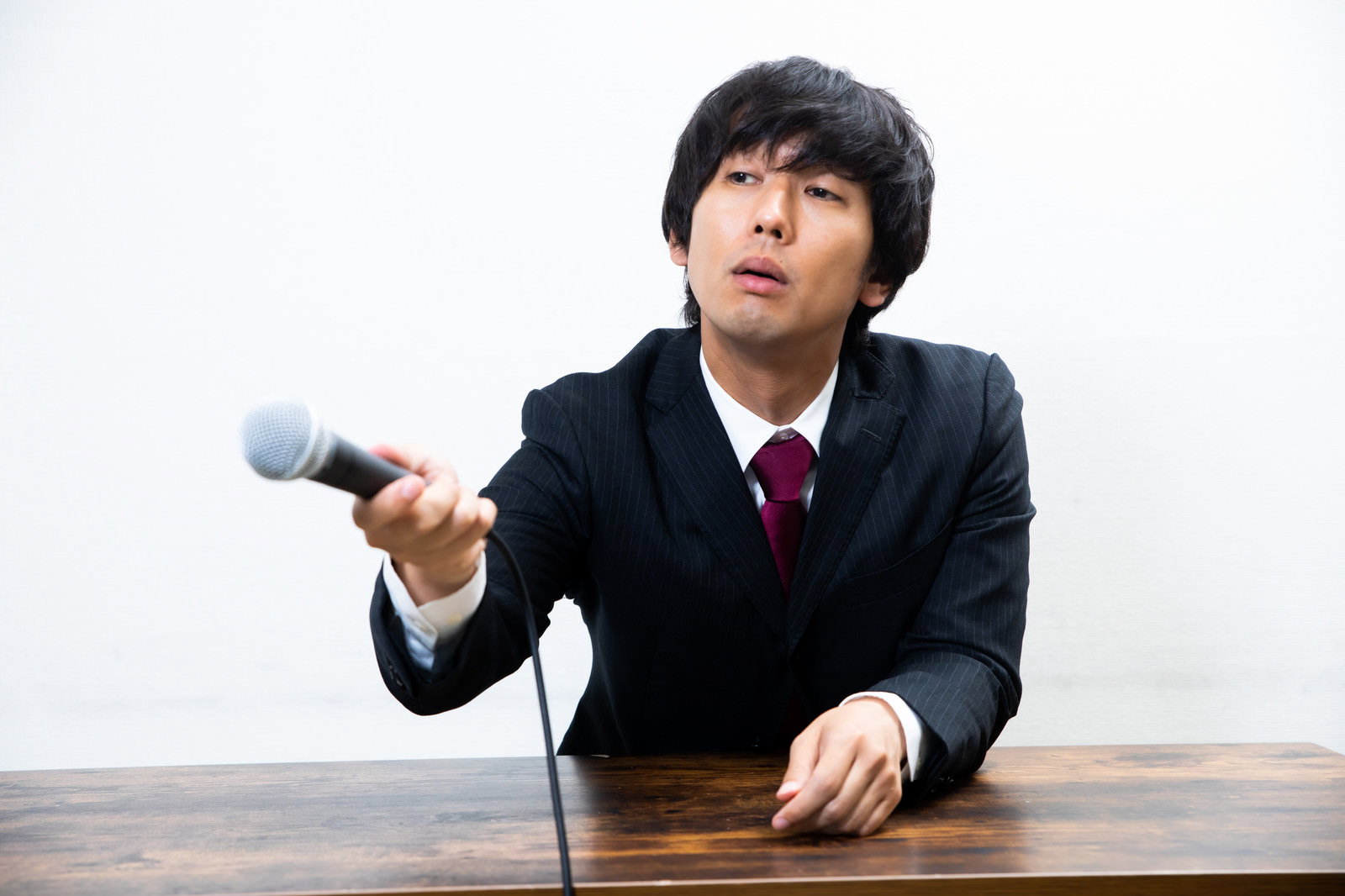記者会見でマイクを持ち呆れた表情を見せるスーツ姿の男性［モデル：大川竜弥］