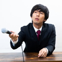 記者会見でマイクを持ちながら呆れて記者に質問をする男性の写真