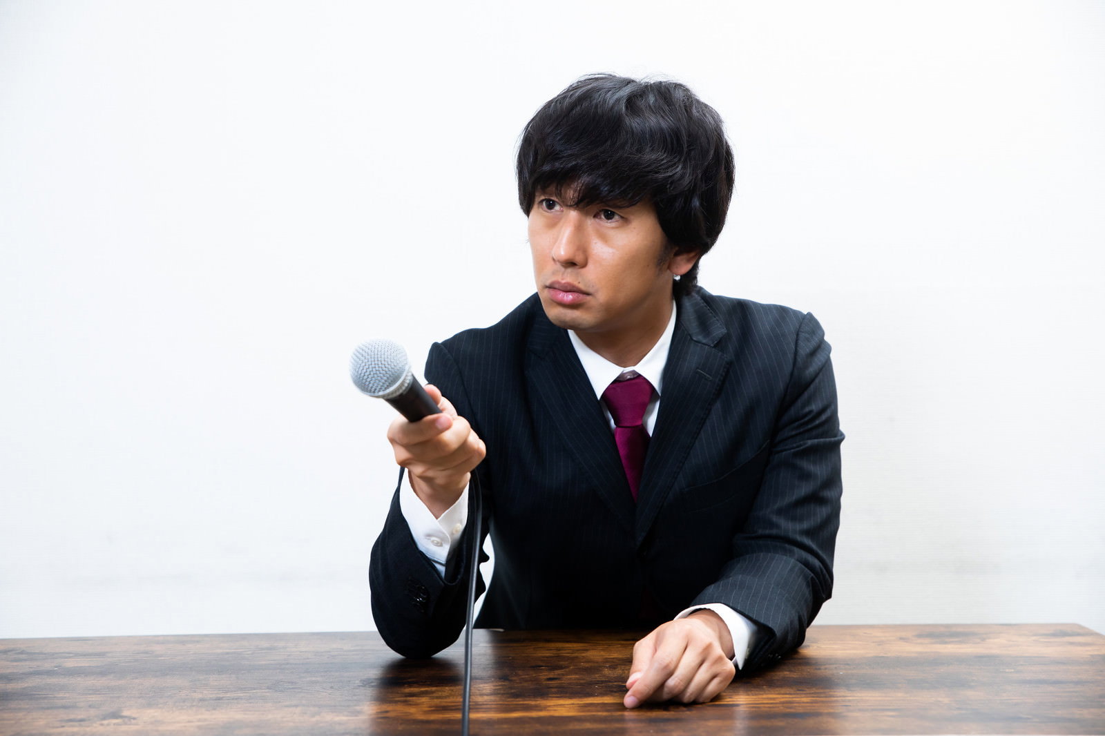 黒いスーツを着た男性記者が険しい表情でマイクを向けている様子［モデル：大川竜弥］