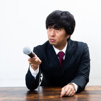 黒いスーツの男性記者が険しい表情でマイクを向ける取材シーンの写真