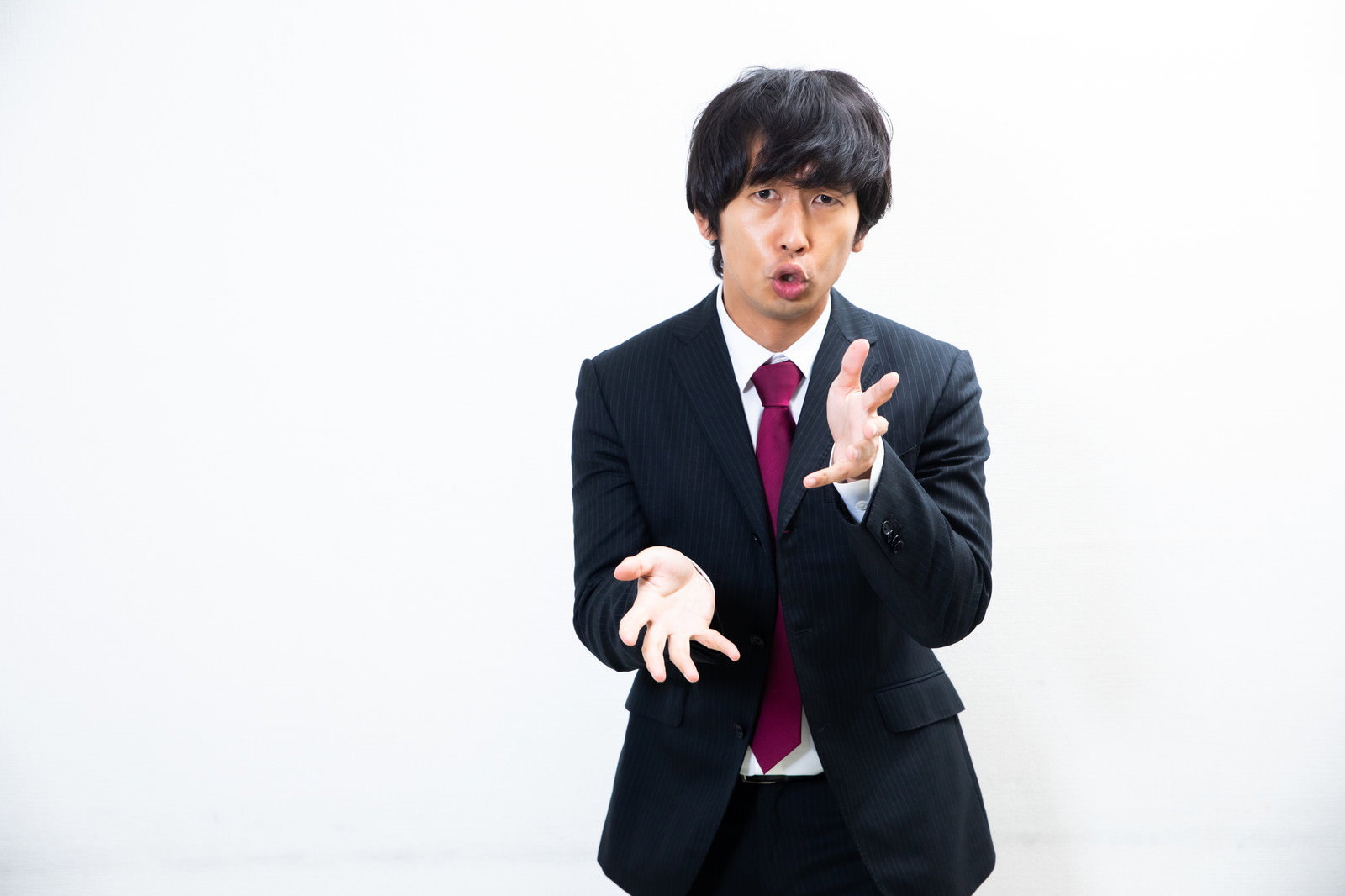 黒いスーツと赤いネクタイを着用した男性会社員が手をくねくねと曲げて説明するポーズ［モデル：大川竜弥］
