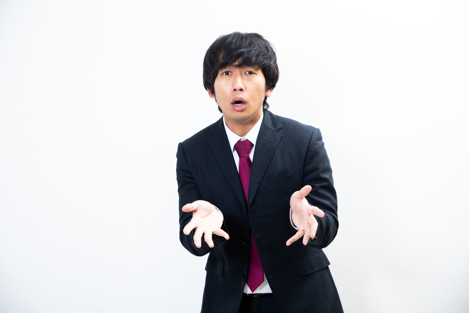 白い背景で両手を広げて説明するスーツ姿の男性［モデル：大川竜弥］