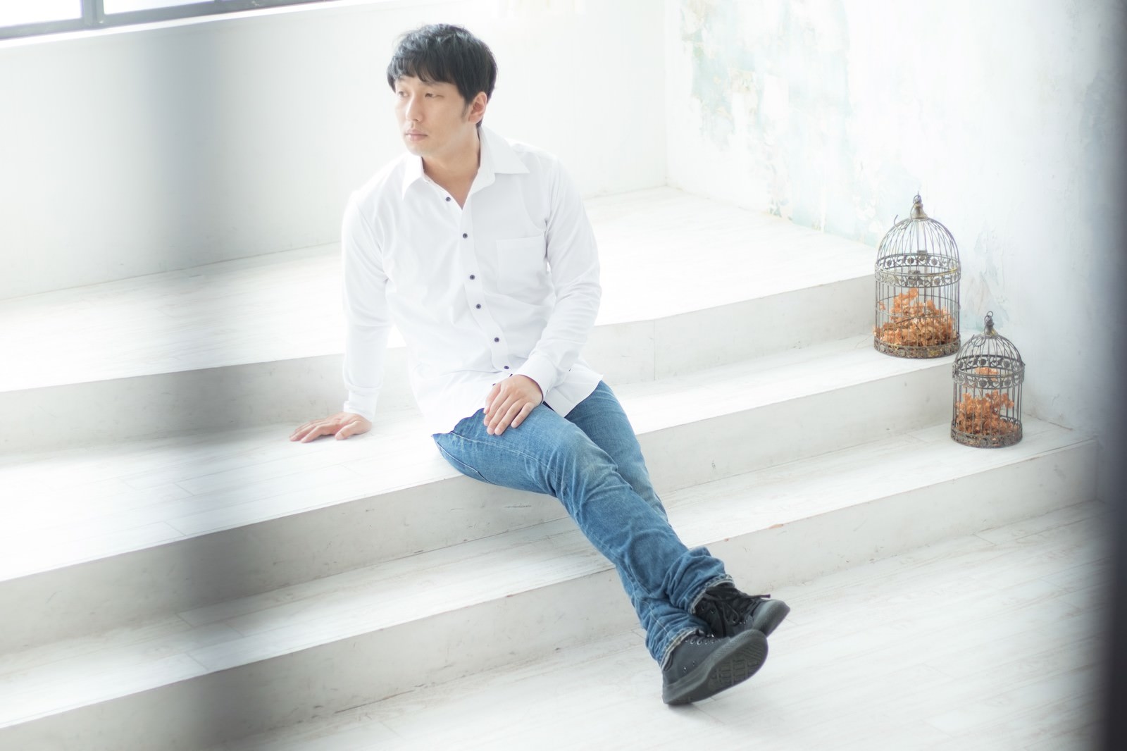 白いシャツとジーンズ姿の男性が白い段差に座っている［モデル：大川竜弥］