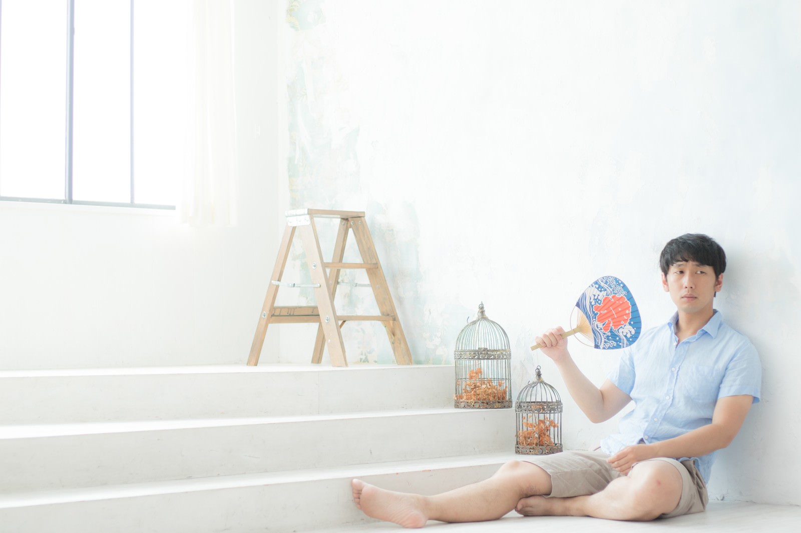 うちわで仰ぎながら白い壁に座る半袖の男性［モデル：大川竜弥］
