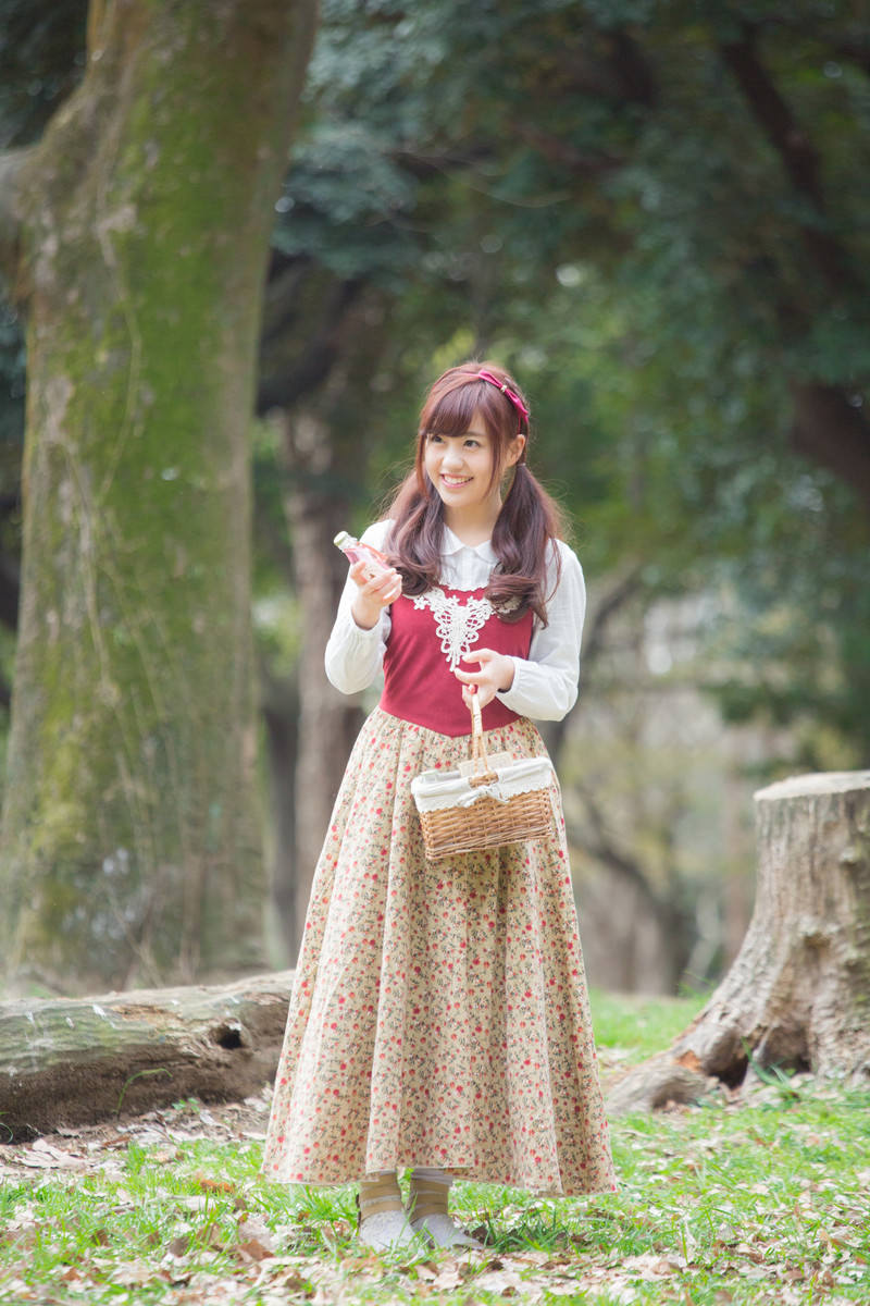 苔むした森の中で、赤いベストと花柄スカート、かごバッグを身につけた女性が小瓶を持ちながら笑顔を見せている［モデル：河村友歌］