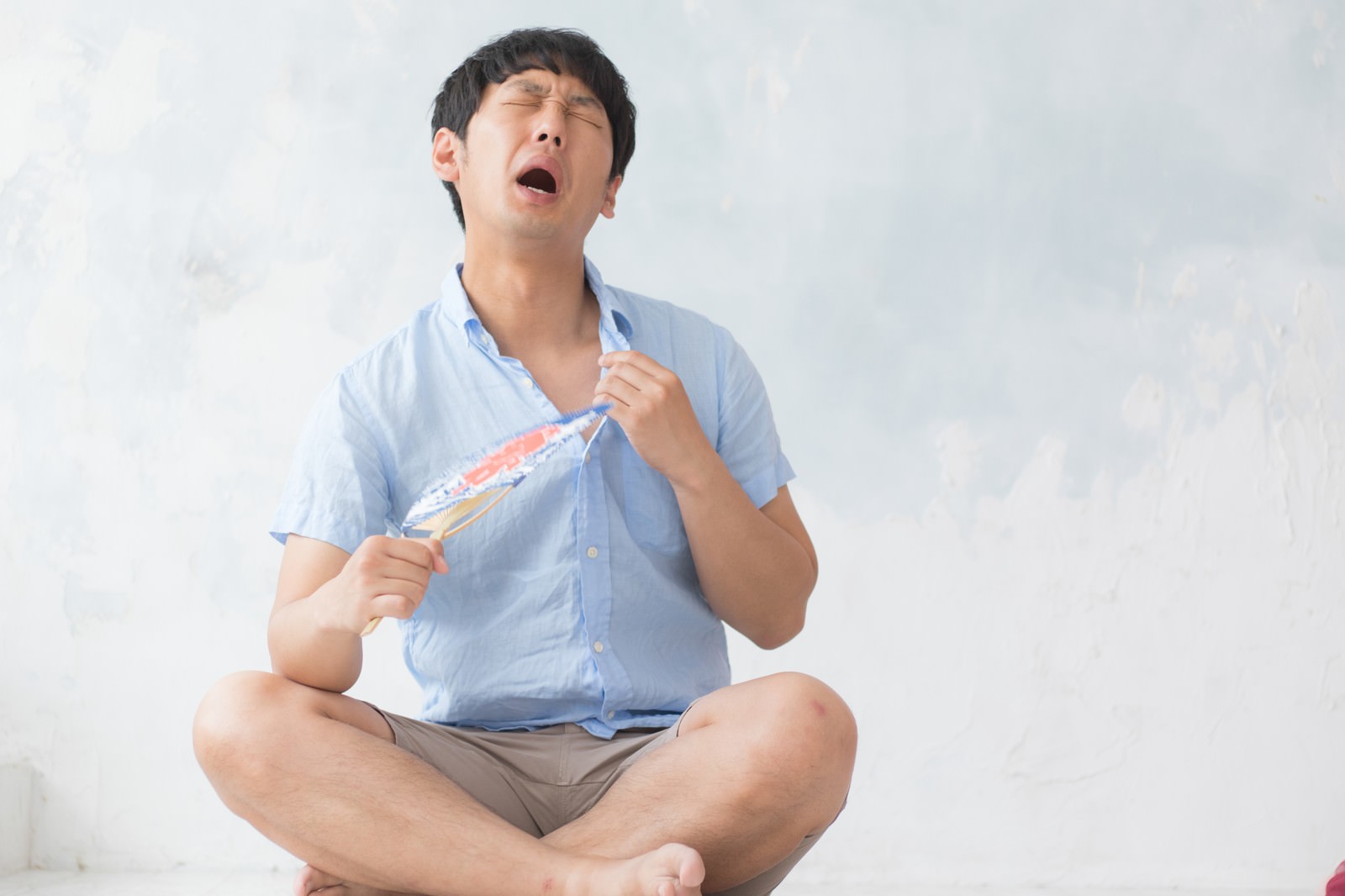うちわで汗をぬぐう男性。水色の半袖シャツ姿で、頭を傾けながら顔の汗を拭いている［モデル：大川竜弥］