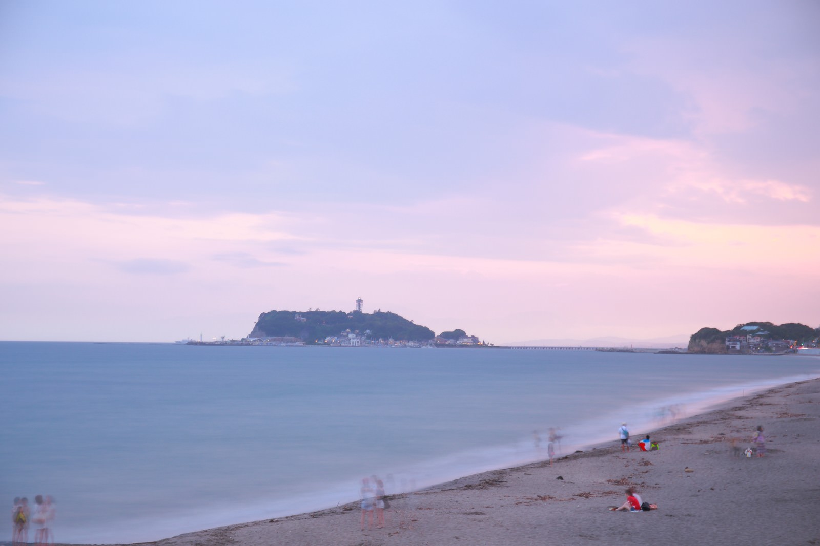 夕暮れ時、薄紫とピンク色の空の下で江ノ島を眺めながら波打ち際で遊ぶ子どもたちと砂浜の人々