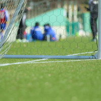サッカーゴールの網越しに見える芝生のフィールドと青い服の選手たちの写真