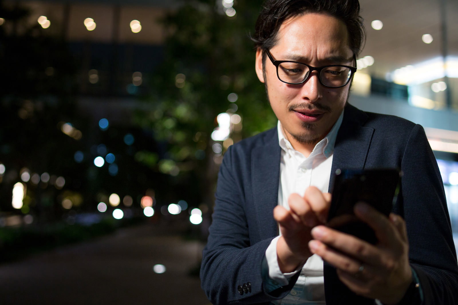 スマートフォンを見ながら検索する眼鏡をかけたスーツ姿の男性［モデル：ゆうせい］