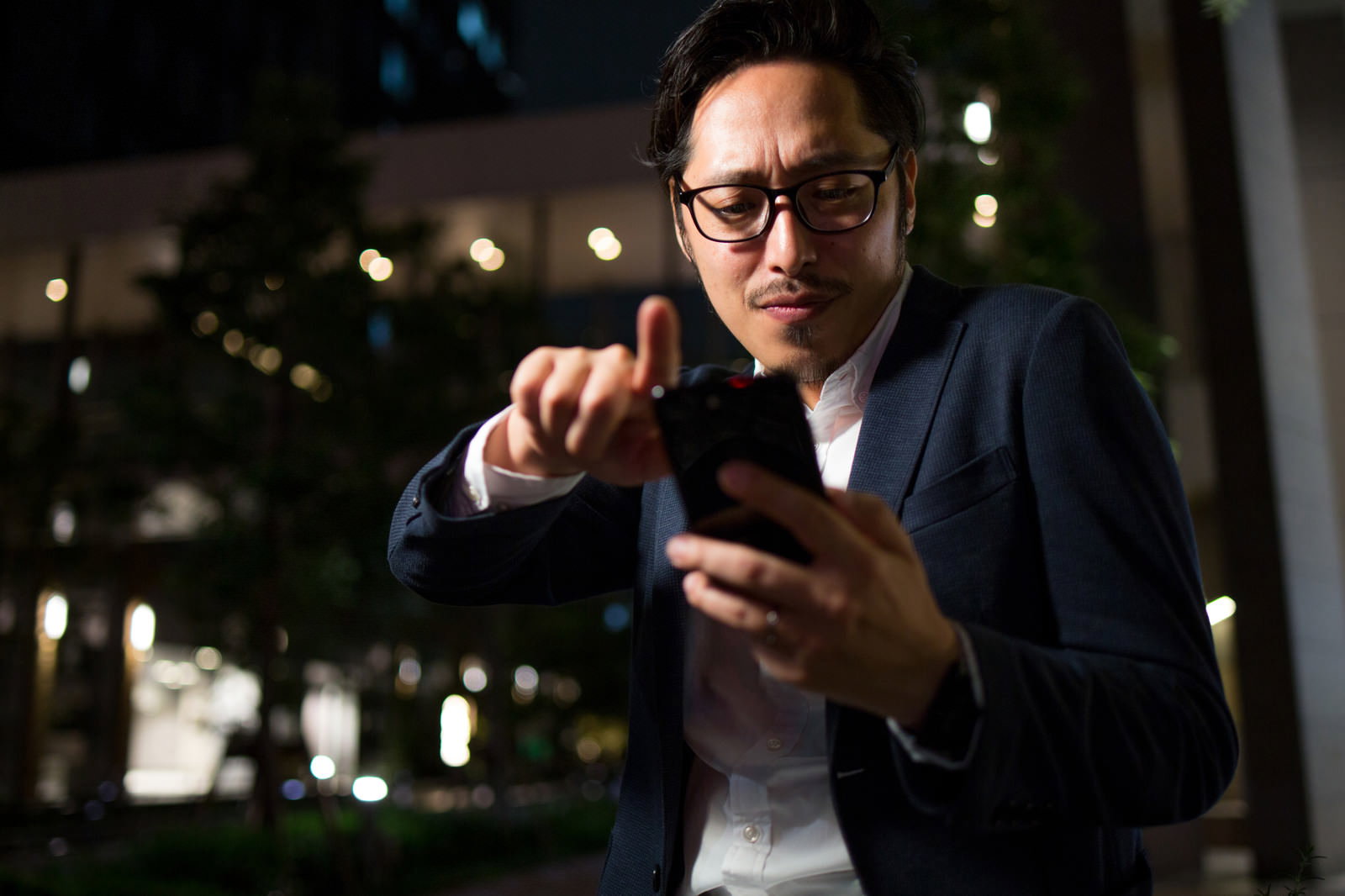 夜間に路上でスマートフォンを操作するスーツ姿の男性［モデル：ゆうせい］