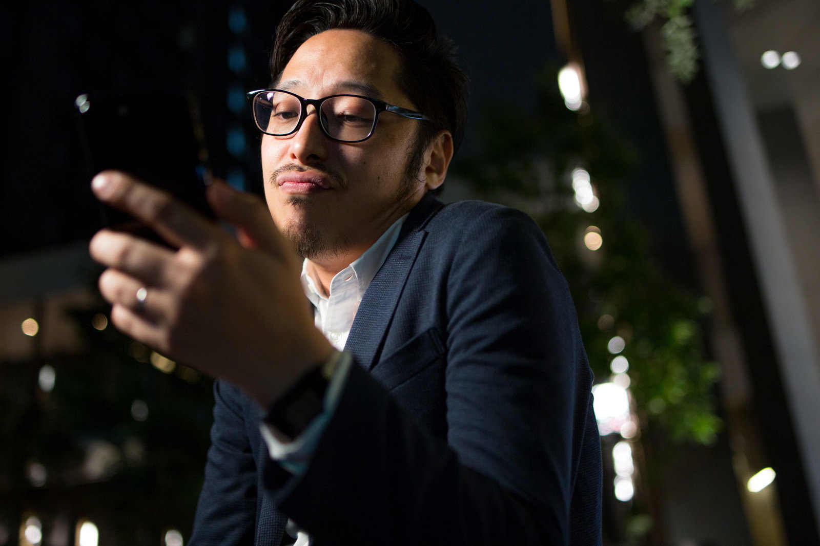 スマートフォンを手に夜間で眉をひそめるスーツ姿の眼鏡男性［モデル：ゆうせい］