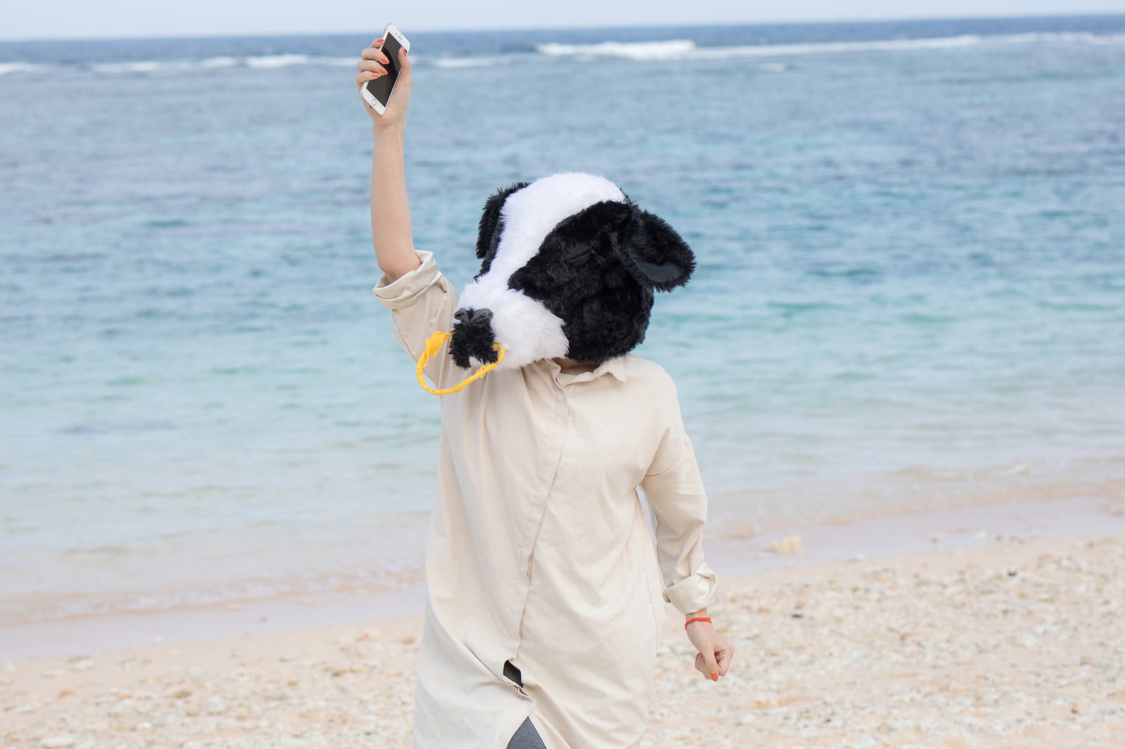 海辺でスマートフォンを掲げて自撮りする女性の後ろ姿［モデル：古性のっち］