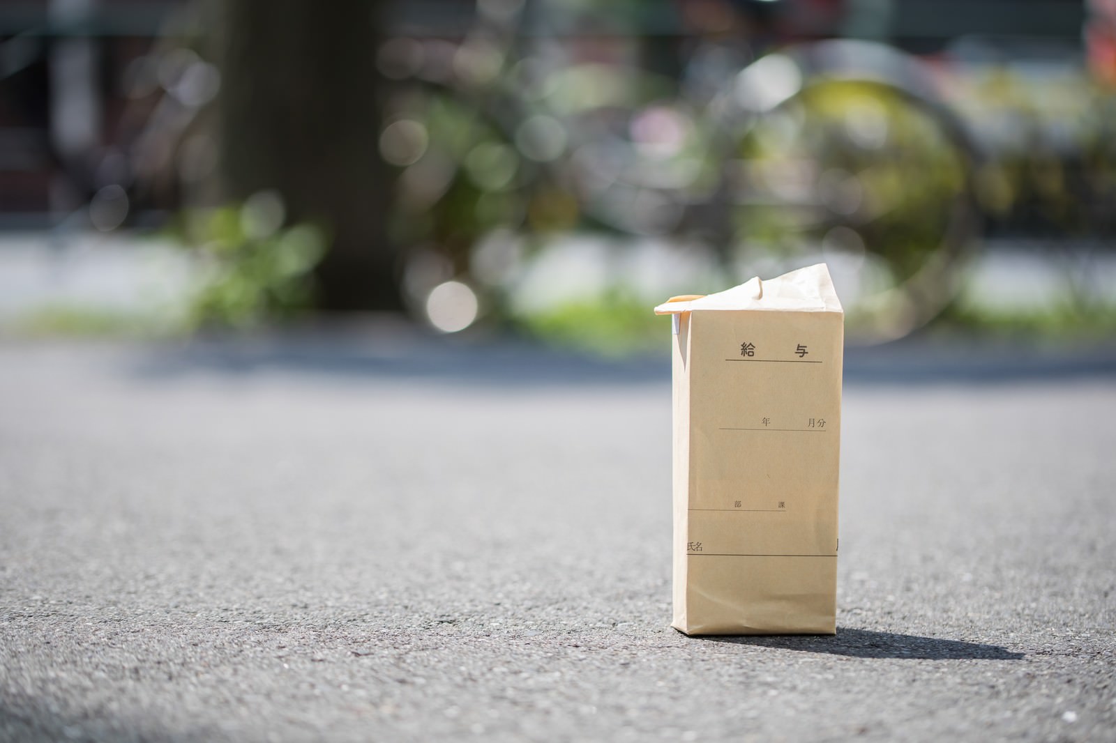 茶色い給料袋が道路上に立てられ、背景に緑ぼけの住宅街がある屋外風景